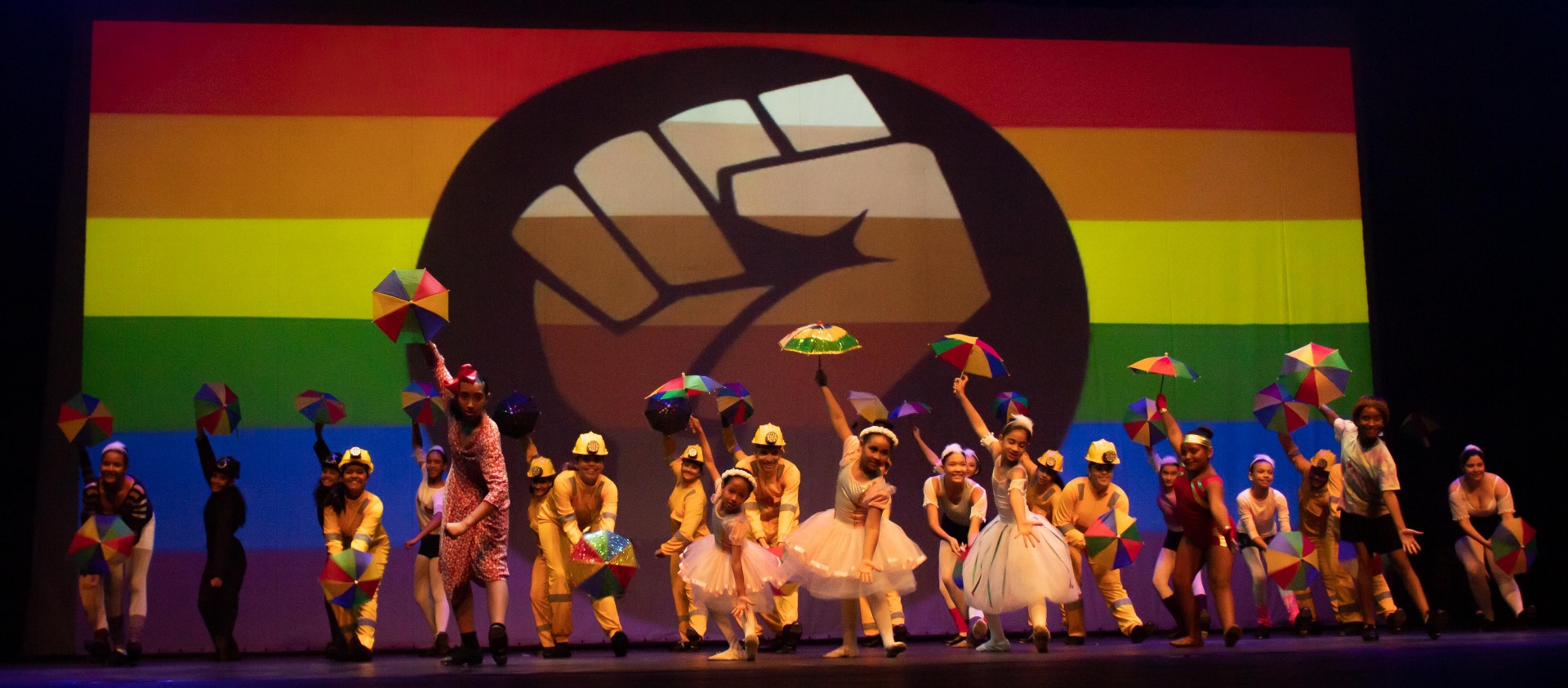 Elenco com sombrinhas coloridas diante de bandeira arco-íris e punho erguido no Billy Elliot