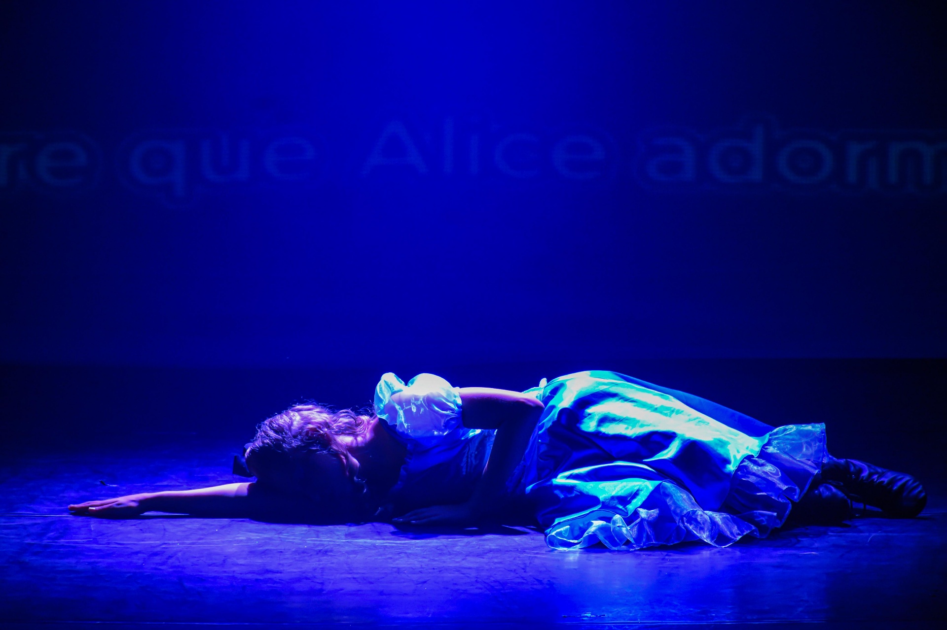Alice adormecida no palco sob luz azul com projeção de texto no espetáculo Alice