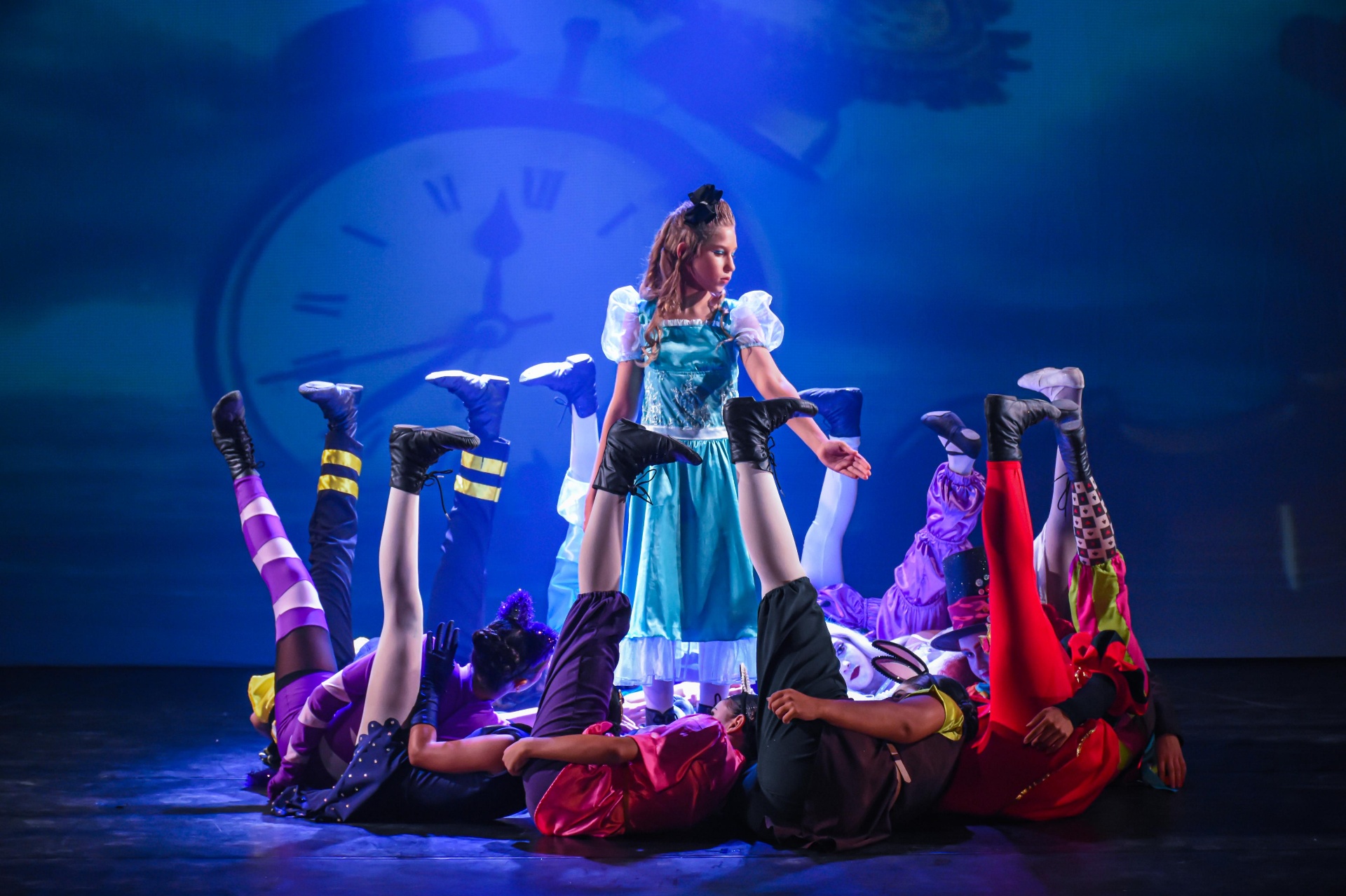 Alice de pé entre pernas erguidas do elenco diante de grande relógio no espetáculo Alice