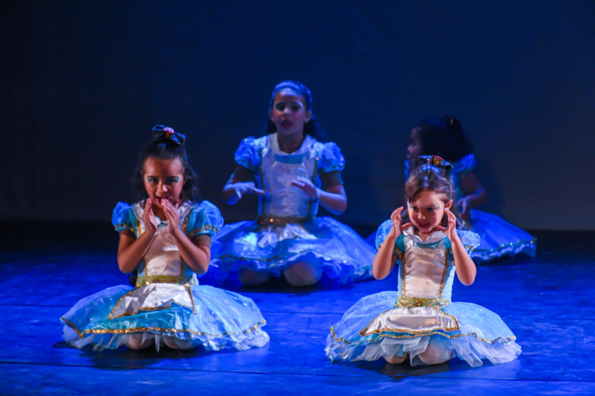 Mini Alices em vestidos azuis cintilantes ajoelhadas no palco do espetáculo Alice