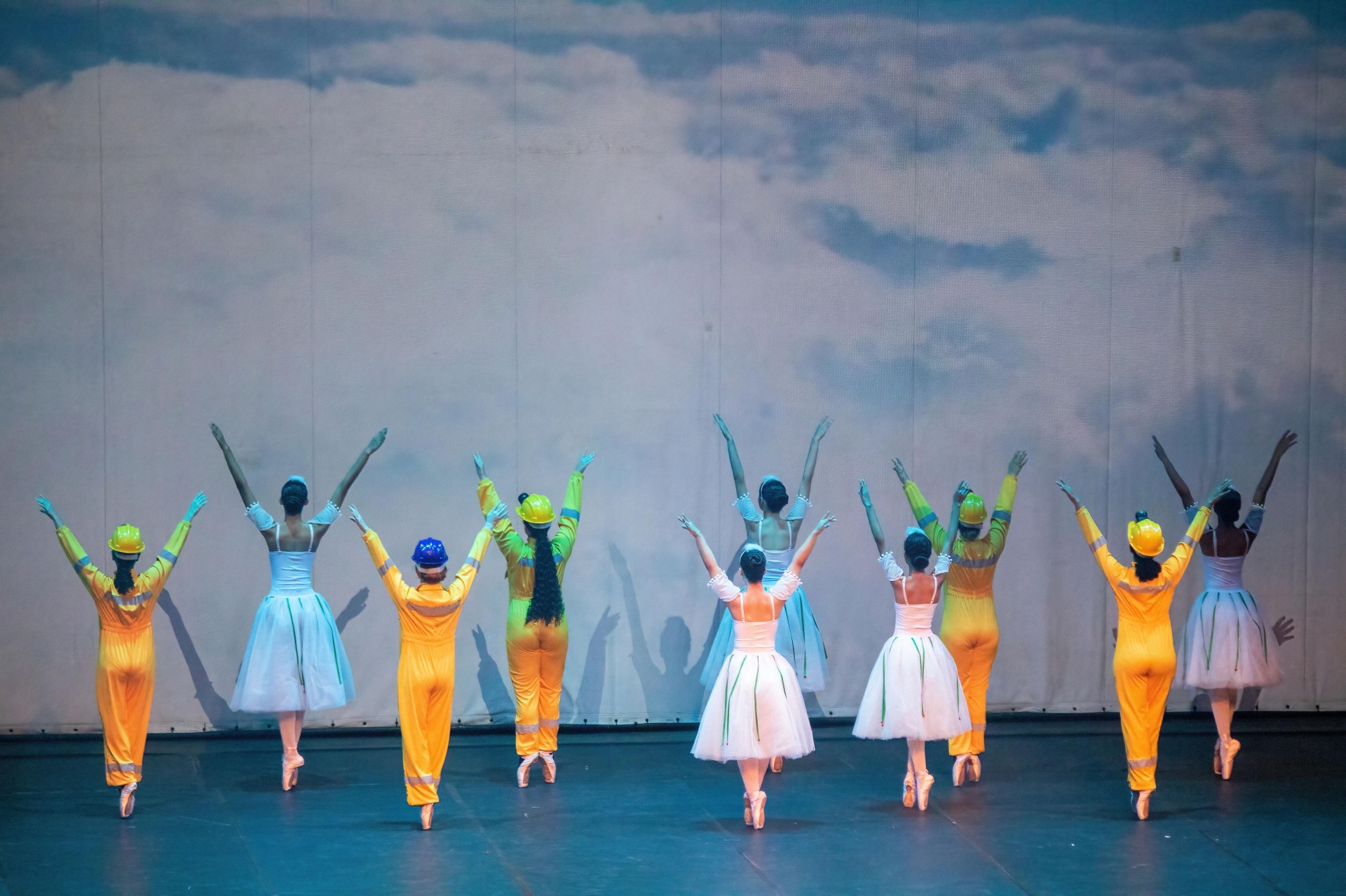 Elenco de bailarinas e mineiros com braços erguidos sob projeção de céu no Billy Elliot