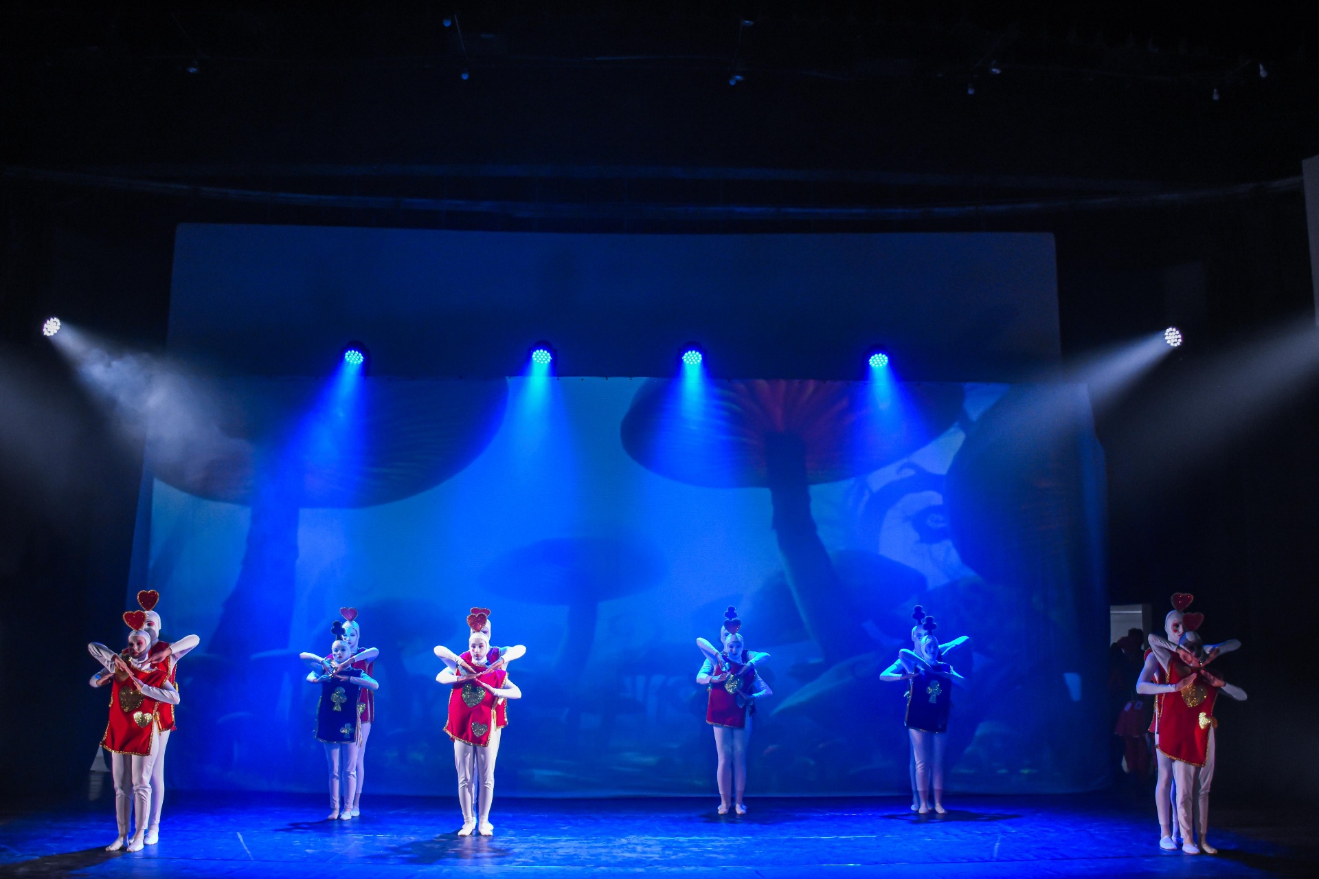 Bailarinas vestidas como peças vermelhas diante de cenário de cogumelos azuis no Alice