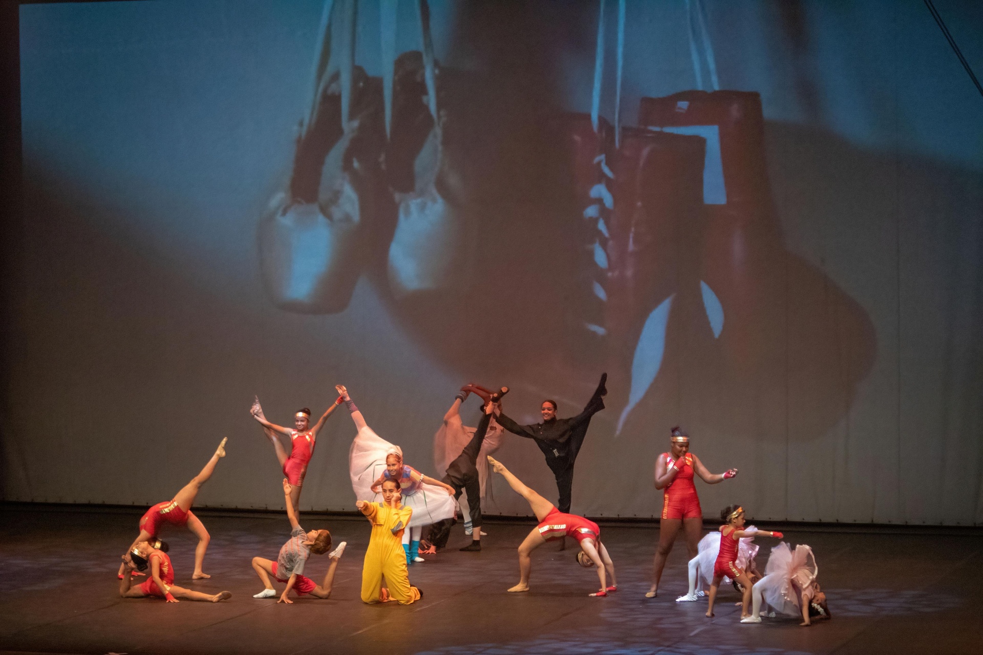 Elenco em acrobacias e poses de dança sob projeção de sapatilhas no Billy Elliot
