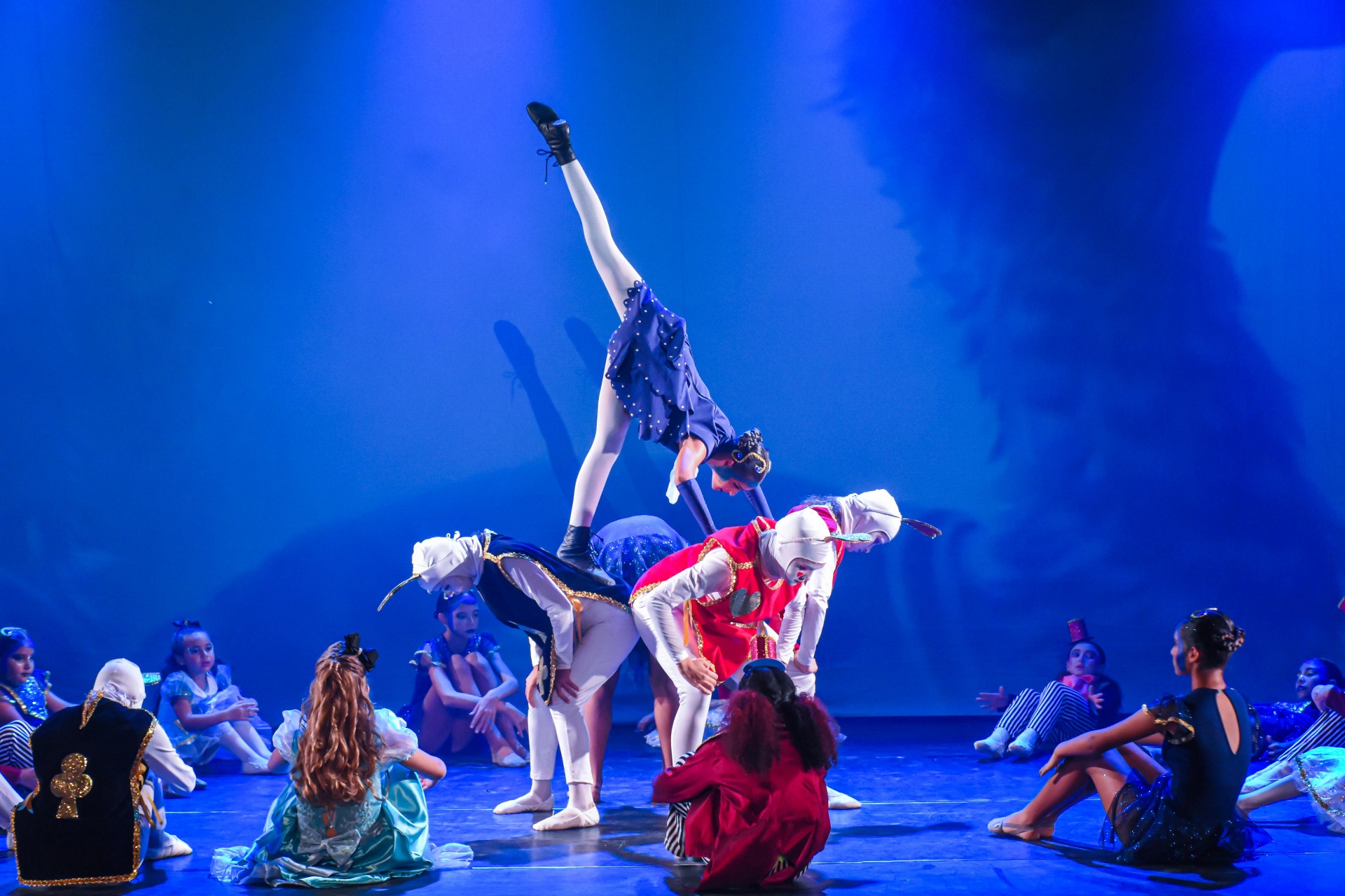 Acrobacia com bailarina em azul sobre elenco em cena de Alice